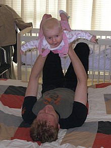 A young father lies on his back on a quilt on the floor. He holds his baby daughter up above him with his arms straight and his hands round her ribcage. The baby has her arms and legs stretched out and arches her back smiling directly at the câmera.