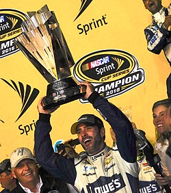 Jimmie Johnson holding a silver trophy in both his hands against a yellow board with sponsors logos