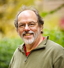 A bearded man in his early sixties grinning, wearing wire rim eyeglasses and an olive drab fleece jacket