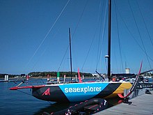 Vu de trois quarts avant, un voilier noir, bleu et jaune amarré au ponton. Bout-dehors et foils rouges. Mât noir.