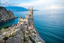 Vogelperspektive auf die Festung, die auf die Klippe gebaut wurde. Die Festung besteht aus hellem Stein und mehreren kleinen Türmen, die auf dem großen runden Turm und dem eckigen Gebäude verteilt sind. Viele Touristen haben sich auf der Plattform vor der Festung versammelt. Im Hintergrund sieht man das Meer und die Felsenküste.