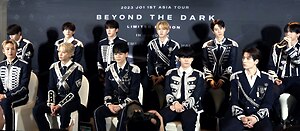 Sebelas orang ahli JO1 duduk dalam dua baris di hadapan latar belakang JO1 1st Asia Tour Beyond the Dark Limited Edition