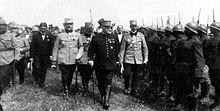 Photographie noir et blanc de trois hommes en uniforme paradant devant des troupes au garde à vous.