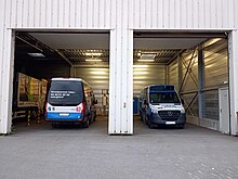 Photographie en couleurs de deux minibus bleus, blancs et noirs. L’un est de dos, tandis que l’autre est de face, et ils sont vus chacun à travers la porte d'un hangar blanc.