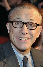 A middle aged man wearing glasses and a grey suit faces forward while smiling.