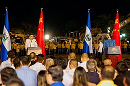 Nayib Bukele and standing at a podium in front of Chinese and Salvadoran flags with a construction crew in the background