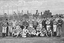 Un groupe d'hommes, habillés en majorité d'un polo sportif, pose en rang pour une photographie. En arrière-plan, des installations sportives.