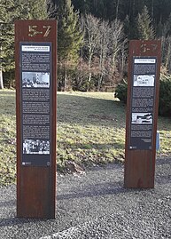 En bordure de route, deux panneaux en bois avec des textes explicatifs et images d'archives.