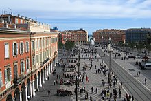 Vue plongeante depuis un immeuble d'une place piétonne traversée par une voie de tramway avec des arcades sur la gauche.