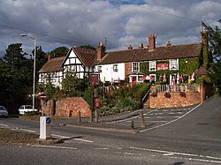 Red brick and tile building on a sloping site with outside raised seating area and car parking to front at lower level adjacent to road down on a sunny day