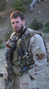 Michael Murphy em camuflagem desértica marrom e bege olhando para a câmera. Ele está vestindo várias peças de equipamento de combate militar verde e segurando uma arma. Há uma colina atrás dele coberta de pedras, sujeira e galhos.