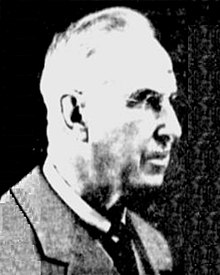 A black-and-white photographic portrait of an older man from the shoulders up, wearing a suit, with a necktie, and eyeglasses