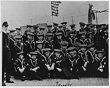 Three rows of men, one seated on the floor and two standing, in a black and white photograph taken on a ship's deck with the American flag flying above