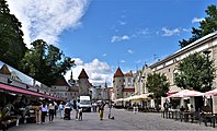 Frontale Farbfotografie eines Marktplatzes mit der Stadtmauer und zwei Wachtürmen, die im Hintergrund sind. Mehrere Außenterrassen stehen an beiden Seiten und der Platz ist belebt.