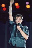 Peter Gabriel bernyanyi dengan sebuah mikrofon, satu tangannya mengepal ke atas.