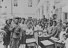 A crowded outdoor space, full of people, mostly young, in civilian clothes; a uniformed soldier with a rifle stands in the middle.