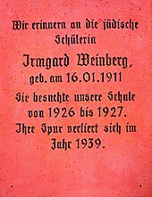 Eine Gedenktafel aus altrosa-farbenem, poliertem Stein, vielleicht Granit. Sie trägt in schwarzer Frakturschrift den Text: „Wir erinnern an die jüdische Schülerin Irmgard Weinberg, geb. am 16.01.1911. Sie besuchte unsere Schule von 1926 bis 1927. Ihre Spur verliert sich im Jahr 1939.“