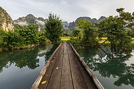 Vue de face d'une passerelle en bois au-dessus d'un lagon, arbres et montagnes karstiques, tôt matin dans les rizières de Vang Vieng.