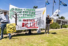 Cloth banner being held up by two people. The banner has a picture of a tree with an arrow pointing to it saying "Carbon capture storage". The banner also has a picture of an industrial facility and an arrow pointing to it saying "Another big lie".