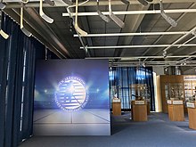 Blue and gray-colored panel in the foreground depicting the hall of fame's logo, with trophies and artifacts on display in the background