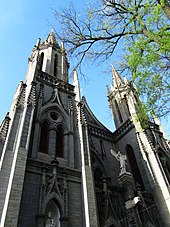 Two ornate spires of grey stone, with a tree branch partially in leaf at right, seen against a blue sky at a sharp upward angle