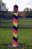 A red/black/yellow striped square-shaped pole with a metal spike on the top side. A metal plaque with an emblem and the words "Deutsche Demokratische Republik" is visible on one face.