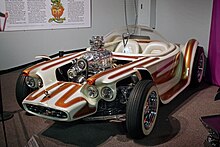 A white car with brown stripes, with open wheels and a clear bubble canopy over twin seats, and exposed, chromed engine with a blower. Beatnik Bandit II and a few of Roth's other cars are also on display in this museum.