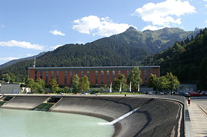 Speichersee Latschau II mit dem dahinter liegenden Lünerseewerk. Es befindet sich gerade im Turbinenbetrieb, zu sehen am Wasseraustritt aus den beiden Öffnungen in der linken Bildhälfte.