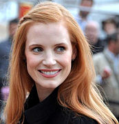 Photographie couleurs au format portrait d'une femme rousse au teint pâle, regardant l'objectif en souriant.