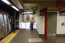 The stairway down to the L train (BMT Canarsie Line) platform at the far south end of the Downtown platform of the 14th Street-Union Square station of the BMT Broadway Line. There is a sign above the staircase, a train at the platform to the left, and and a white wall to the right.