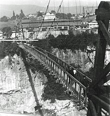 Photo en noir et blanc prise depuis le versant côté Haute-Savoie et montrant la passerelle piétonne suspendue reliant les deux rives. Les pylônes supportant les câbles et le tablier de la passerelle sont en bois. Quelques personnes sont en train de traverser.