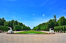 Frontale Farbfotografie einer langen Rasenfläche, die bis zum Horizont führt und von zwei Wegen mit Baumalleen begrenzt ist. Hinter den Alleen sind im Hintergrund größere Bäume. Gestutzte Hecken und zwei Hirschskulpturen, die Wasserfontänen in ein kleines Wasserbecken schießen, sind an der linken und rechten Vorderseite. Am vorderen Bildrand ist ein breiter Kiesweg.