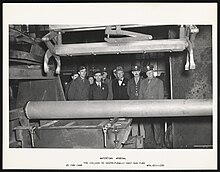 A photograph of the 100,000th centrifugally cast gun tube being produced inside a large factory warehouse with Army officials watching in the background. The photo caption reads “WATERTOWN ARSENAL The 100,000th Centrifugally Cast Gun Tube 20 FEB 1944.”