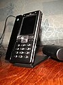 A candybar style phone on a desk stand that is placed on top of a wooden desk. A cable leads from the stand off-frame.