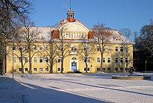 Es ist Winter, der Platz vor dem Schulgebäude und der flache, kreisrunde Brunnen sind schneebedeckt, die Laubbäume vor dem Museum sind kahl. Vor uns steht quer ein dreigeschossiges, symmetrisches Gebäude mit sandgelber Fassade und rotem Walmdach. In der Mitte springt ein drei Fenster breiter Gebäudeteil etwas hervor. Unten führt dort ein kurzes Treppchen zur blau gestrichenen Eingangstür, über der eine Laterne die Stufen beleuchtet. Auf dem Dach über diesem Gebäudeteil wird die Außenwand noch um ein weiteres Stockwerk nach oben erweitert und von einem eigenen, kleineren Walmdach gekrönt (sogenanntes „Zwerchhaus“). Dieser höchste Teil der Fassade mündet oben in einen flachen Rundbogen, unter dem über einem Fensterband eine große Uhr zu sehen ist. Diese ist rechts und links von dekorativen Ornamenten (weiß auf gelbem Grund) umgeben. Oben auf dem First des Zwerchhauses thront ein achteckiges Türmchen mit einer Fahnenstange.