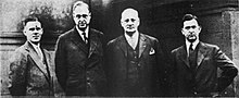 A black-and-white photograph of four men in suits, standing side-by-side. A middle-aged Costello poses with his hands behind his back.