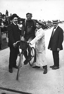 Photographie noir et blanc d'un cycliste entouré de quelques personnes debout.