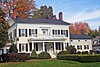 A large white two and a half story house with an open porch supported by four columns, topped by a balustrade to form a balcony