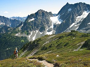 Photograph of snowy mountain peaks with a hiker in the bottom-left atop grassy terrain