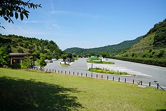 Eine helle, flache Betonfläche in einer grünen hügeligen Landschaft, im Hintergrund sind bewaldete Berge zu erkennen. Die Betonfläche weitet sich von der Zufahrt rechts unten auf, sodass mehrere parallele Fahrstraßen entstehen. Auf der Betonfläche sind schräge, rechteckige Parkplatzmarkierungen in weißer Farbe zu erkennen. Die Parkplatzfläche wird von kleinen Grüninseln mit Bäumen und Gräsern unterbrochen. Am linken Rand der Fläche ist das Dach eines kleinen Gebäudes zu sehen.