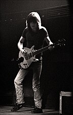 This photo is in black and white. Malcolm, aged 29, is shown in left profile, playing guitar with both hands. He has over shoulder-length hair, and is wearing a dark T-shirt, jeans and shoes.