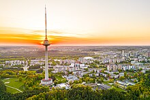 Farbfotografie von einem hohen Fernsehturm mit Sonnenuntergang. Vorne sind Grünanlagen und im Hintergrund viele Häuser.