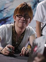 A brown-haired man in his forties, wearing glasses and a white top, signs an autograph at a convention.