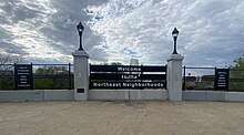 Black sign with white text says "Welcome to the Northeast neighborhoods", on a fence, surrounded by lamp posts. Smaller black signs with white text to either side list neighborhoods: Logan Park, St. Anthony East, Marshall Terrace, Holland, Bottineau, Sheridan, St. Anthony West, Northeast Park, Beltrami, Waite Park, Windom Park, Audubon Park, Columbia Park.