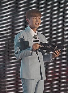 Leeteuk standing in front of a LED display, shuffling cue cards and holding a microphone in his right hand