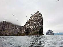 A large, triangular rock rises from the misty waters, with more islands behind and Northern Gannets flying around it.