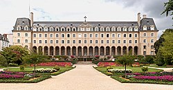 A large four-storey sand-coloured building with 14 arches at the ground-floor entrance and a large landscaped formal garden in front. The photograph was taken during the summer, so many flowers are in bloom and there are leaves on the bushes and trees.