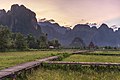 Passerelle en bois élevée sur pilotis et envahie de feuilles de Nelumbo nucifera (Lotus sacré), menant vers une hutte au toit de paille, en face des montagnes karstiques, au coucher du Soleil, à Vieng Tara Villa, Vang Vieng, durant la mousson.