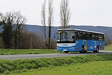 Photographie en couleurs d’un autocar bleu, blanc et noir sur une route bordée par des prés verts. En arrière-plan se détachent quelques arbres, et le relief montagneux.