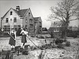 Auf dem alten Bild von 1946 benutzen zwei junge Frauen jeweils eine Hacke bei der Gartenarbeit im Gemüsebeet. Im Hintergrund steht das Schulgebäude.